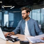 A man financier, auditor sits in the office . he holds documents, checks accounts. types on a laptop